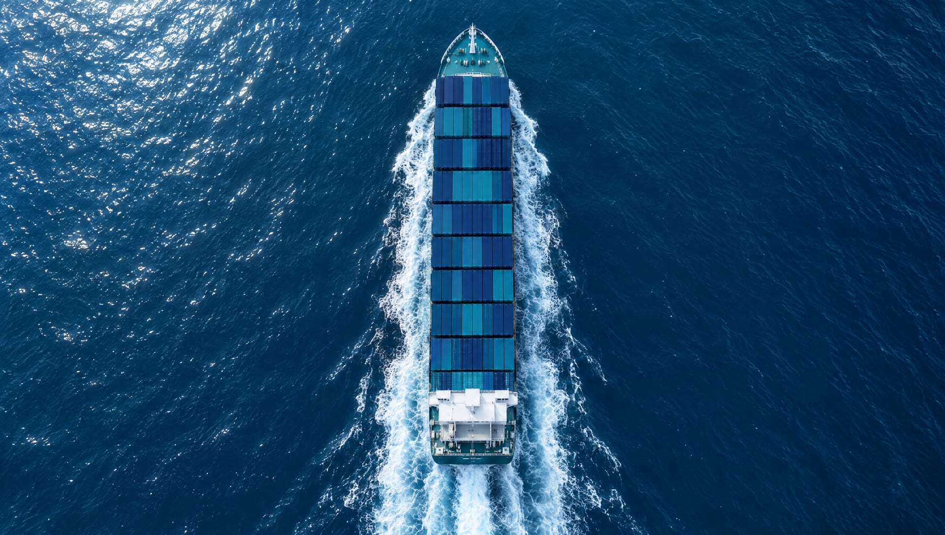 A container ship seen from directly above, cutting through deep blue ocean with a white wake